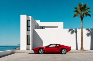 Red vintage sports car beside a white modernist villa, with palm shadows, blue sky and the sea beyond.