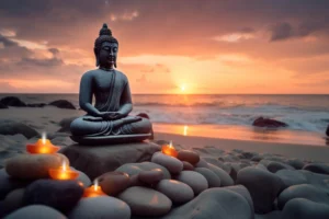 Stone Buddha statue surrounded by smooth pebbles and glowing candles on a beach at an orange ocean sunset.
