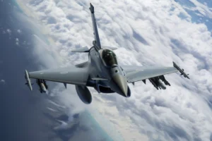 Modern fighter jet banking above a vast white cloud deck in a cool-toned aerial scene.