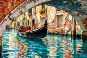 Black gondola gliding beneath an old brick bridge on a bright Venetian canal, surrounded by weathered buildings and turquoise reflections.